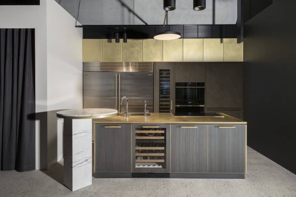Modern kitchen with dark wood cupboards, built-in stainless steel appliances, marble-topped side table, wine cooler, and minimalist lighting fittings, set against a polished concrete floor and dark feature walls.