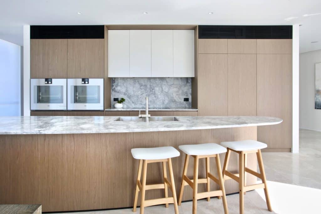 Modern kitchen with light wood cabinets, marble worktops, built-in ovens, and three white bar stools at a large island. The space is bright, minimalistic, and features a neutral colour palette.