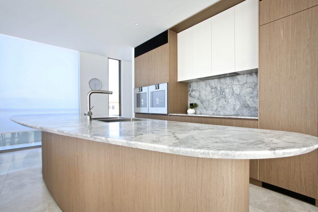 Modern kitchen with light wood cupboards, a large marble island with an integrated sink, built-in ovens, and a marble splashback; natural light streams in from a tall window.