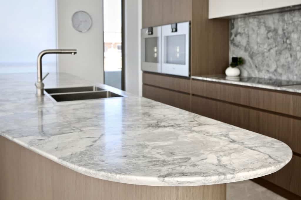 Modern kitchen with a marble worktop, built-in sink and tap, light wood cupboards, two wall ovens, and a marble splashback. A small plant decorates the counter near the ovens.