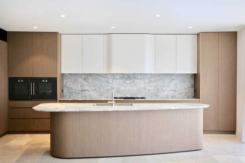 Modern kitchen with a large marble island, wooden cupboards, white upper cupboards, built-in ovens on the left, and a marble splashback. The space is bright and minimalistic with recessed ceiling lights.