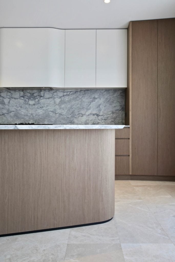Modern kitchen with light wood cupboards, white upper cupboards, and a grey marble splashback. The worktops are white with a subtle pattern, and the floor is covered in light beige tiles.