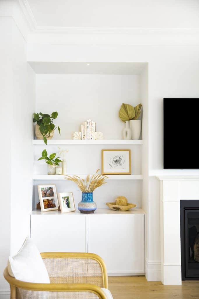 A white living room bookcase displays plants, framed photos, books, and decorative objects. A rattan chair is in the foreground, and a TV is partially visible on the right above a fireplace. The decor is modern and minimal.