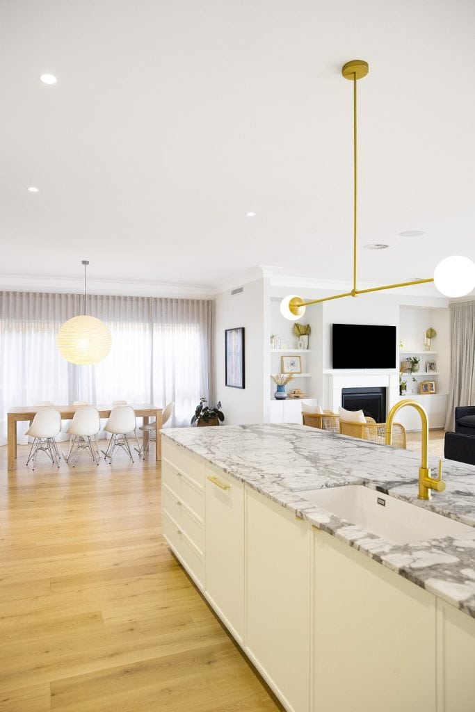 Bright, modern kitchen with a marble island and gold tap, open to a living area with a TV, shelves, and a dining space featuring a wooden table and white chairs, all with light wood floors and large windows with sheer curtains.