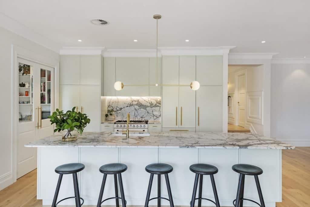 Modern kitchen with a marble island, five black stools, minimalist white cupboards, gold accents, built-in oven, marble splashback, pendant lights, and a potted plant on the worktop.