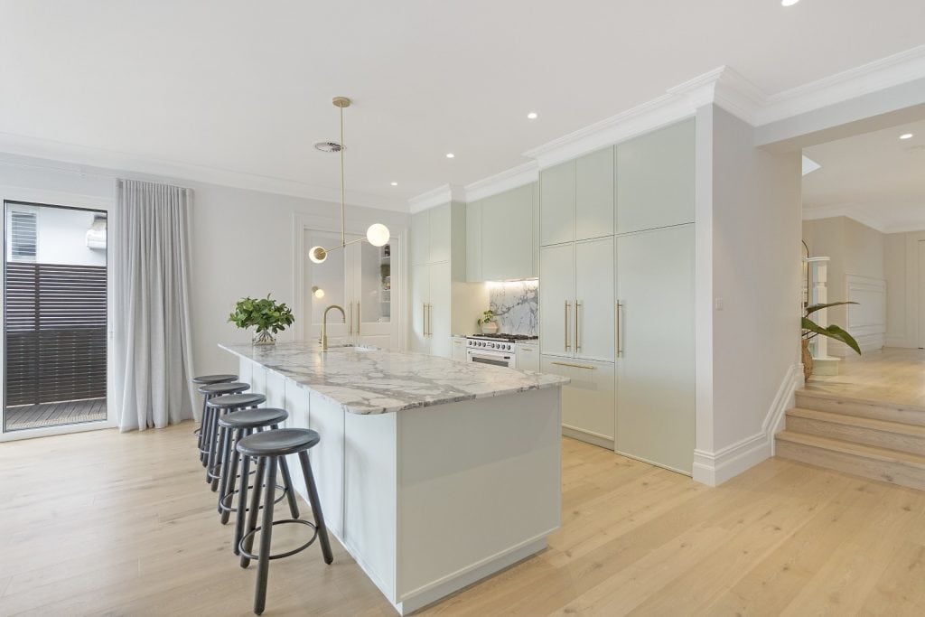 Modern kitchen with a marble island, four black stools, pastel cupboards, gold fittings, and light wood floors. Large windows with sheer curtains let in natural light. Stairs and open hallway are visible in the background.