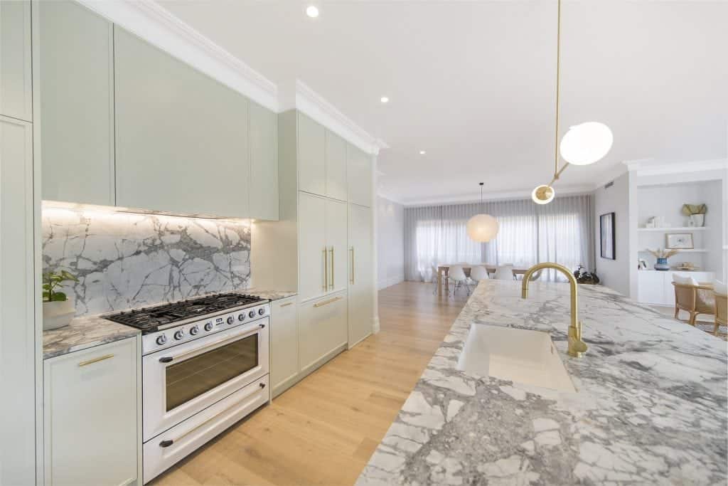 Modern kitchen with light green cupboards, marble worktops and splashback, gas cooker, built-in fridge, brass fittings, pendant lights, and an open view into a bright dining and living area with wooden flooring.