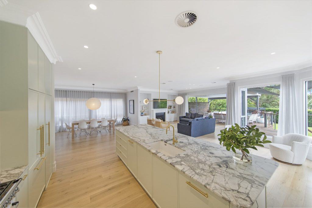 Modern open-plan kitchen and living area with marble island, wooden floors, large windows, dining table, white armchair, and sliding doors opening to a patio and greenery outside.