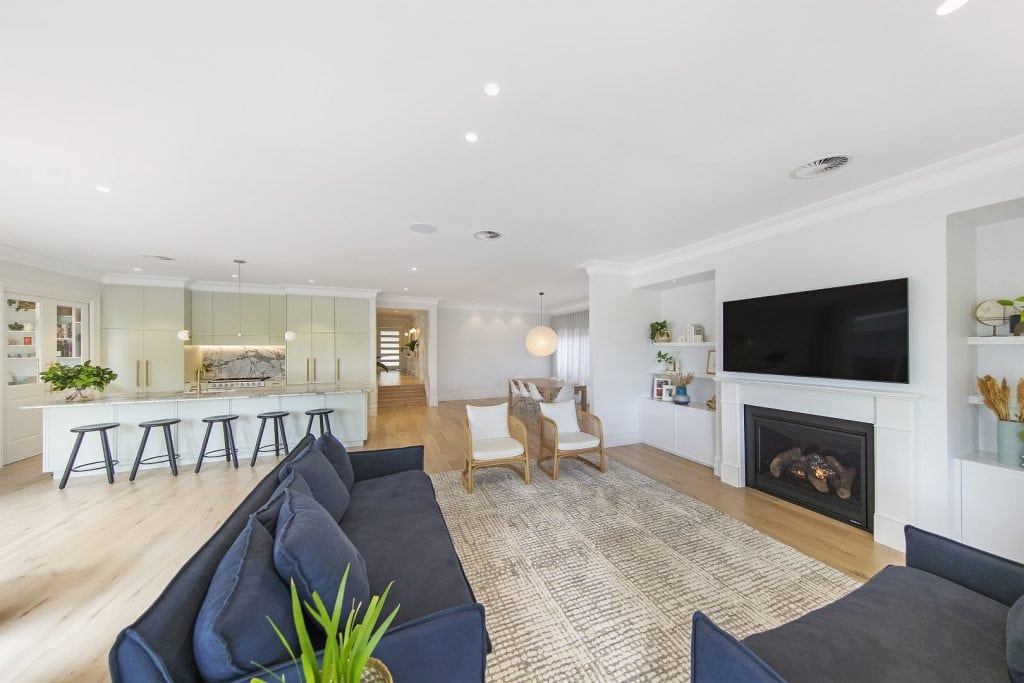 A modern, open-plan living space with a kitchen, island seating, and a living area. The room features a large TV above a fireplace, blue sofas, armchairs, and light wood flooring. The space is bright and airy with minimalist decor.