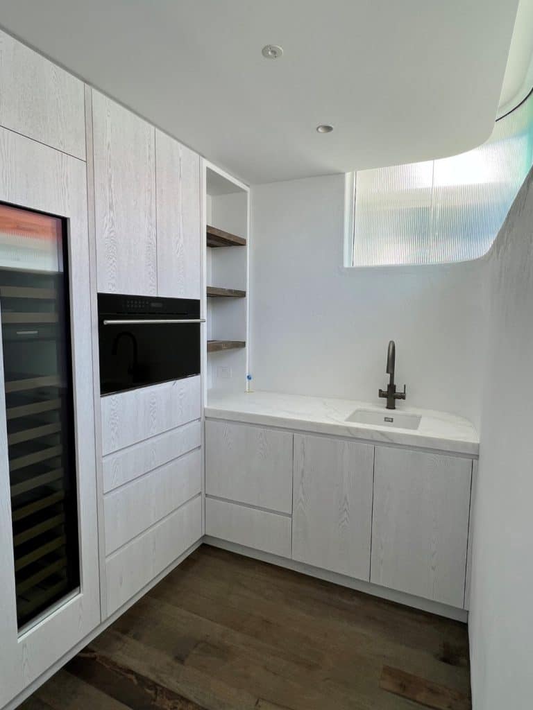 Minimalist modern kitchen with white wood cupboards, built-in black oven, wine cooler, open shelves, and a white worktop with an under-mounted sink and black tap, set against light walls and dark wood flooring.