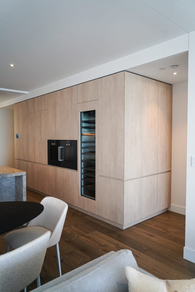 Modern kitchen with light wood cabinetry, a built-in black oven, and a tall glass-fronted wine fridge. The space features minimalist design with clean lines and a cosy dining area with beige chairs.