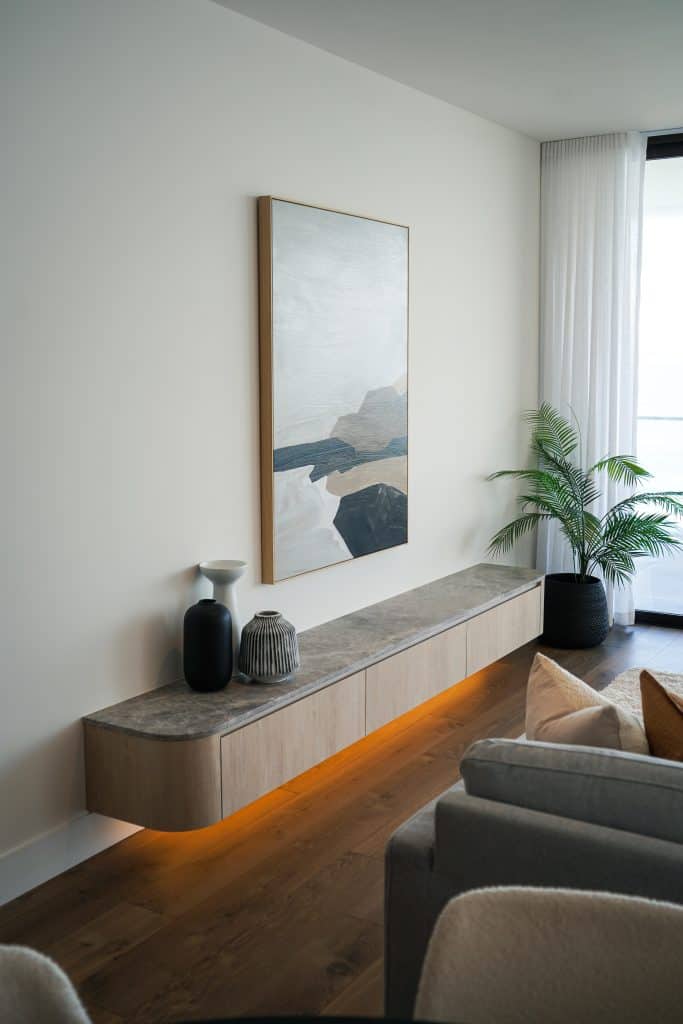 A modern living room features a floating wooden sideboard with a stone top, decorated with two vases and a jar. Above it hangs abstract art; a potted plant and floor-to-ceiling curtains are nearby. Warm lighting glows under the sideboard.