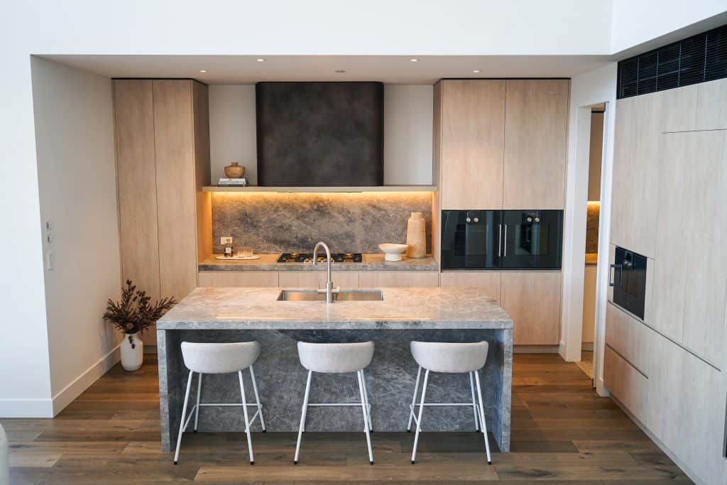 Modern kitchen with a grey marble island and three stools, light wood cupboards, built-in ovens, and under-cabinet lighting. A vase with dried flowers sits on the floor near the island.