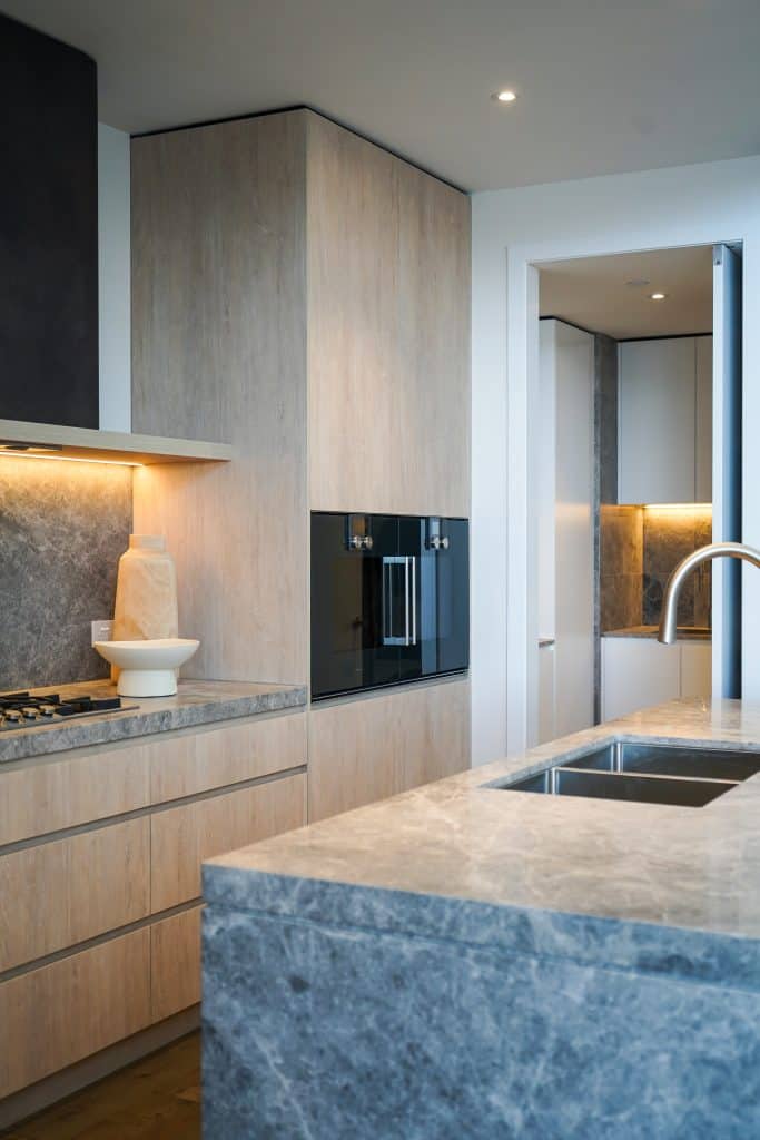 Modern kitchen with light wood cupboards, grey marble worktops, built-in oven, gas hob, under-cupboard lighting, a sink on a marble island, and minimalistic decor in a bright, clean space.