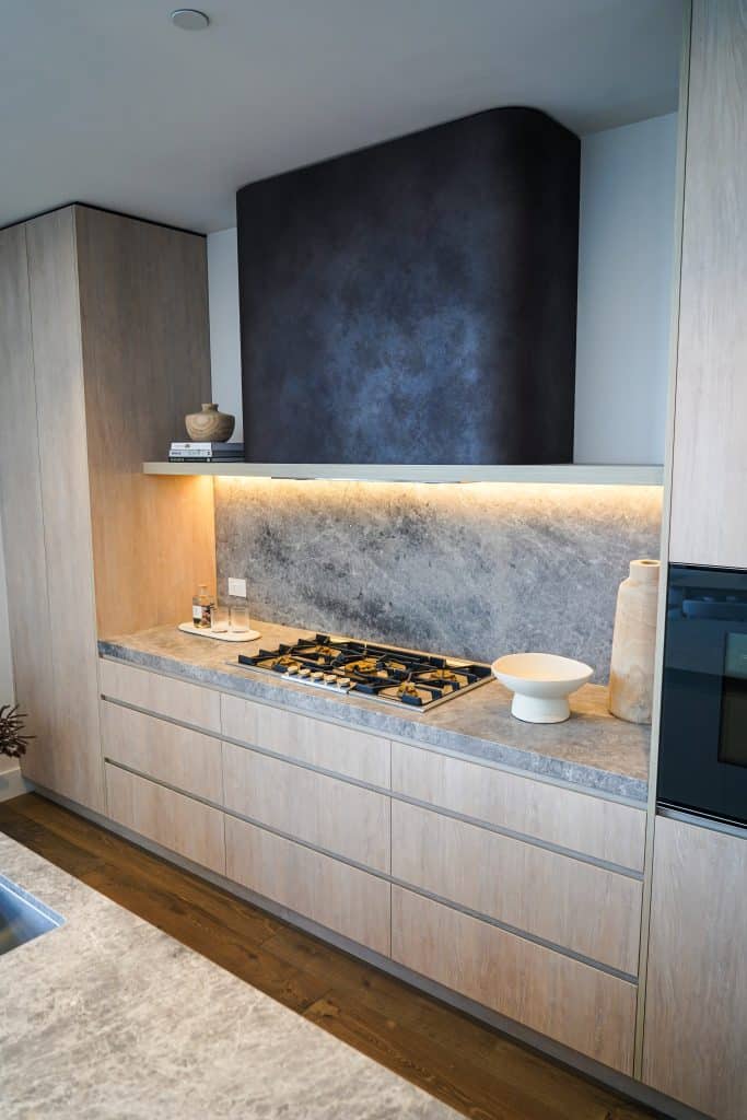 Modern kitchen with light wood cabinets, grey stone worktops and splashback, a built-in gas hob, under-cabinet lighting, minimal decor, and a dark extractor hood above the cooker.