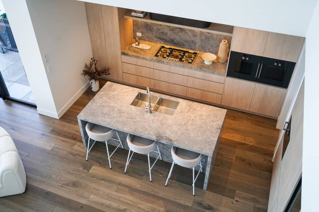 Modern kitchen with light wood cabinets, a grey stone island with a sink, four bar stools, built-in hob, and wall ovens. The space features wooden floors and minimalist decor.