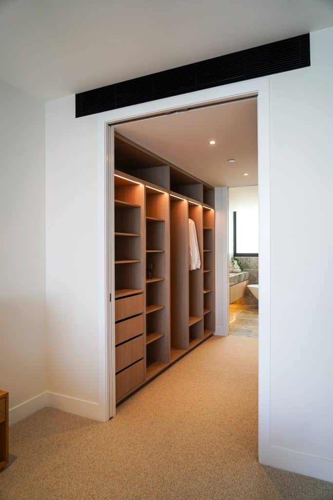 A modern walk-in wardrobe with open wooden shelves and drawers, built-in lighting, and hanging space with clothing. The wardrobe leads to a bathroom with a large window and bath.