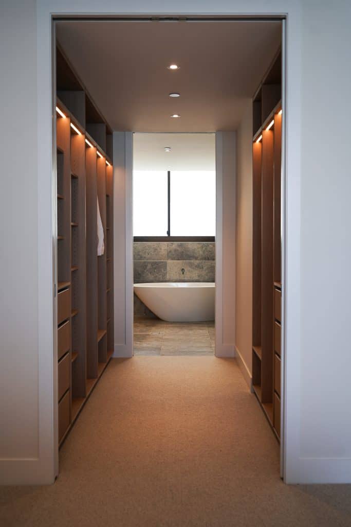 A modern walk-in wardrobe with wooden shelves and drawers on both sides, leading to a bathroom with a freestanding white bath under a large window.