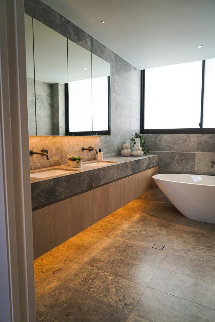 Modern bathroom with grey stone tiles, a floating vanity with under-cabinet lighting, double basins, large mirrors, decorative plants, and a white freestanding bath near a large frosted window.