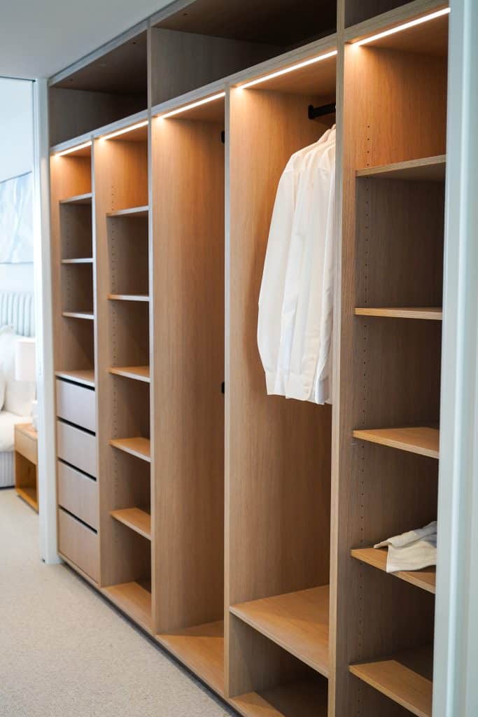 A modern wooden wardrobe with open shelves, drawers, and hanging space. Only a few white shirts are hanging, and one white garment is folded on a lower shelf. The wardrobe is neatly organised and minimally filled.