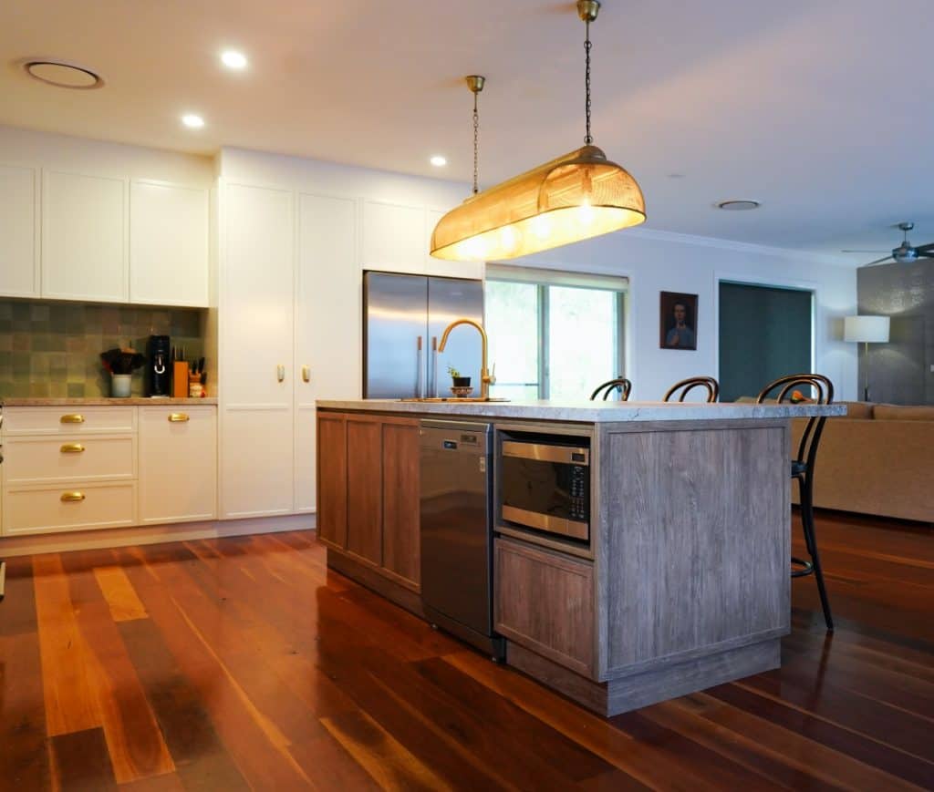 Modern kitchen with wooden floors, a large island featuring a built-in microwave and dishwasher, pendant lighting overhead, white cupboards, stainless steel appliances, and a view into a living room area.