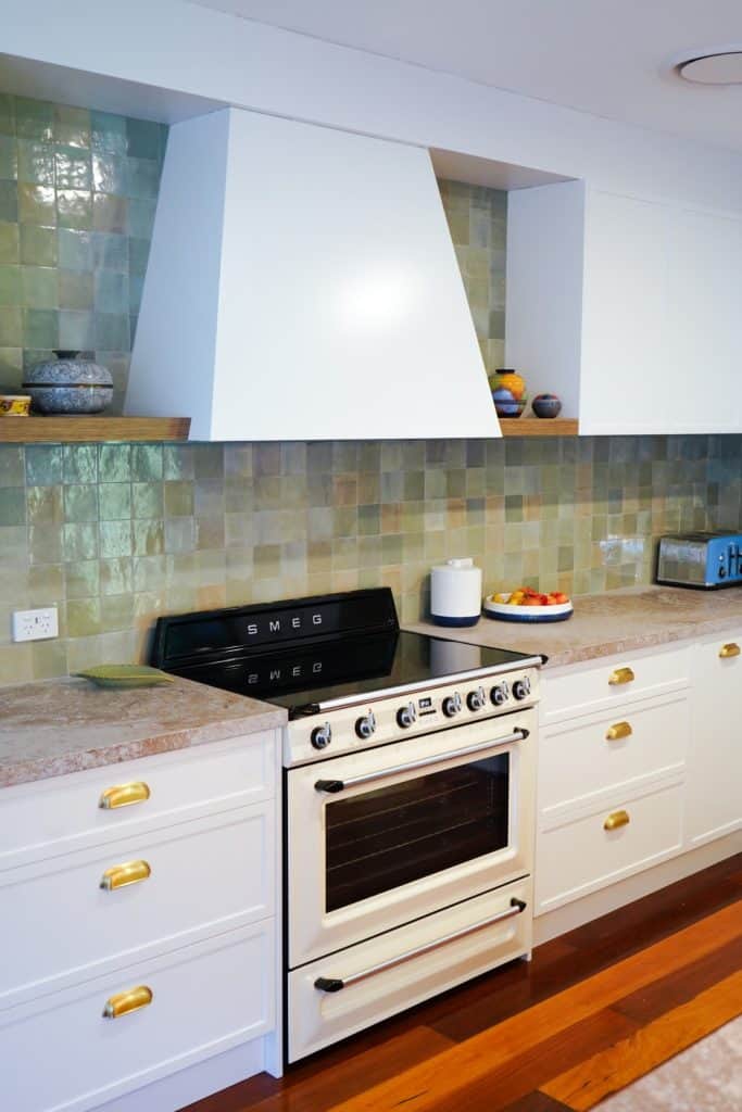 A modern kitchen with a white SMEG oven, glossy green tile splashback, white cupboards with gold handles, and light stone worktops. Decorative bowls and a fruit plate are on the counter.