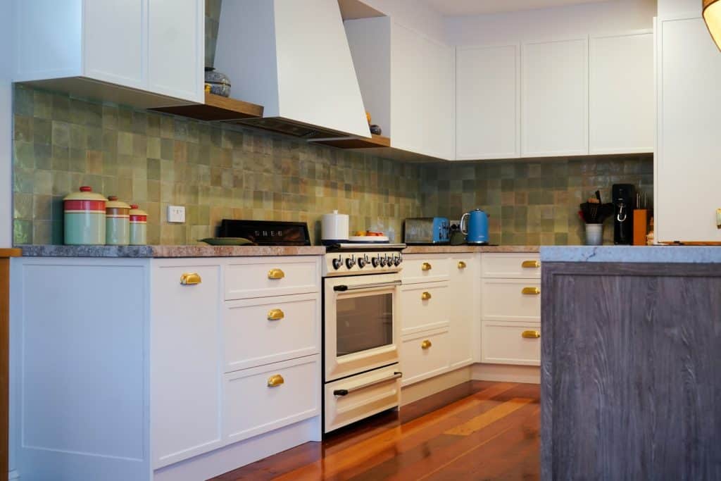 Modern kitchen with white cupboards, gold handles, a gas cooker, green tile splashback, and wooden floors. Worktops have jars, a kettle, a coffee maker, and other small appliances.