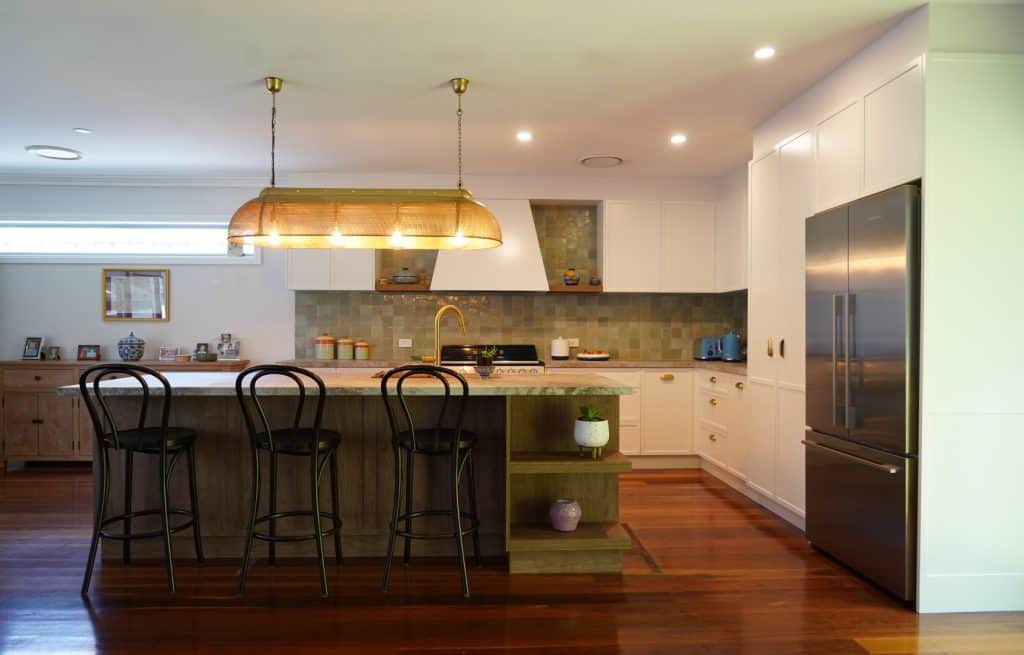 Modern kitchen with a large island, three black bar stools, stainless steel fridge, pendant light, wooden floors, white cupboards, and green tiled splashback. Decorative items and kitchenware are on the counters and shelves.