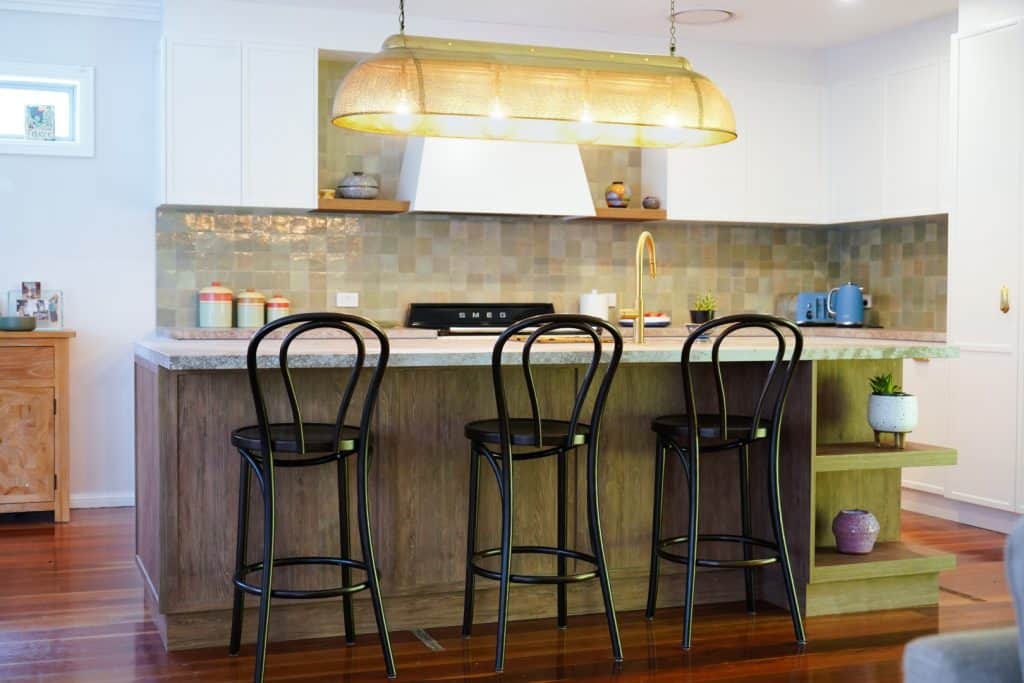 Modern kitchen with a wooden island, three black bar stools, pendant lighting, tiled splashback, and various small appliances and decor on the counter. The floor is wooden, and the space appears bright and inviting.