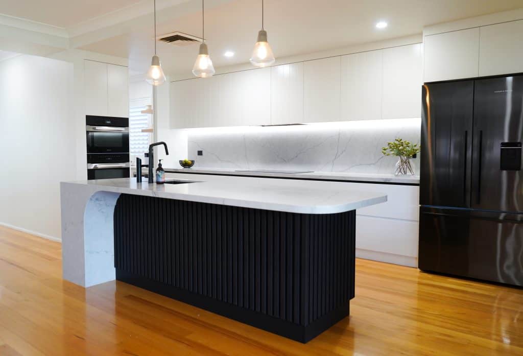 Modern kitchen with white cabinetry, marble splashback, a black fridge, built-in ovens, pendant lights, and a central island featuring a white worktop and black textured base. Wooden floors reflect ambient lighting.