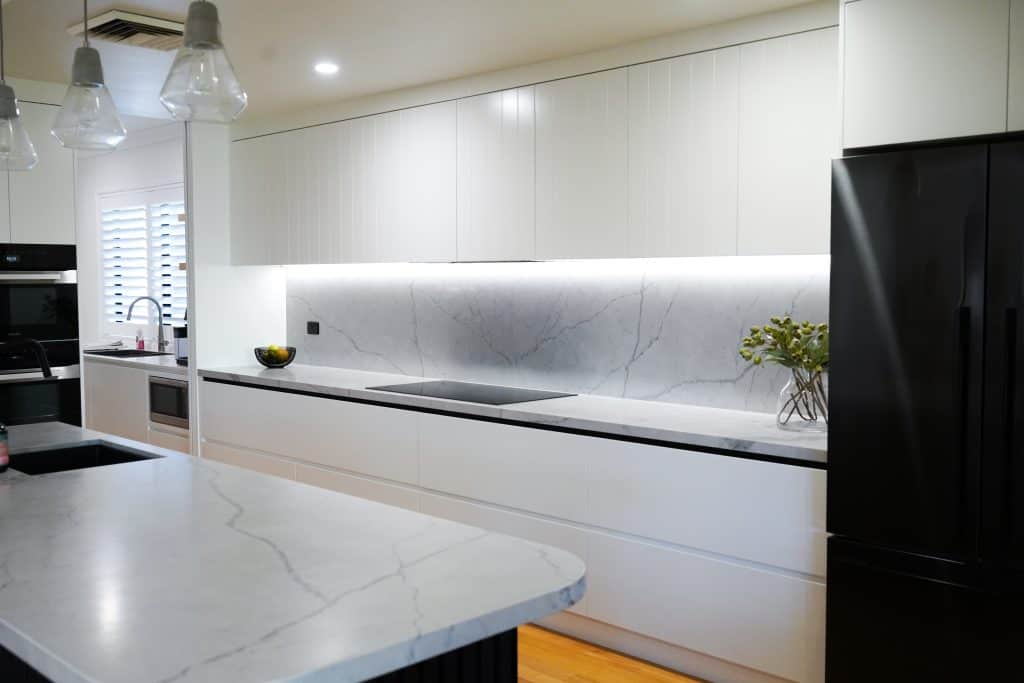 Modern kitchen with white cupboards, marble worktops and splashback, black appliances, an island, and pendant lights. A small vase with greenery sits on the counter near a black fridge.