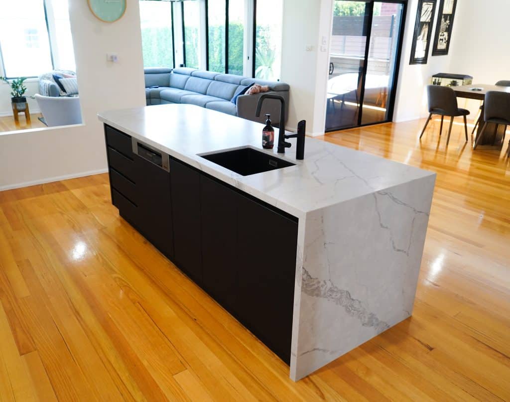 Modern kitchen with a marble island featuring a black sink and tap, set on polished wooden floors. A grey sectional sofa is in the background near large windows and sliding glass doors.