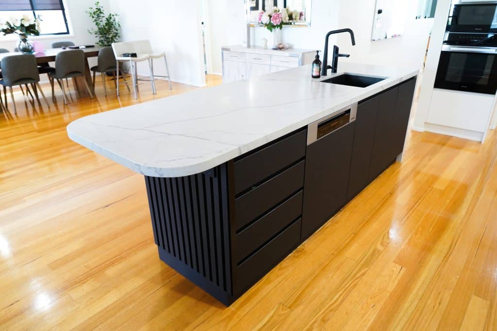 A modern kitchen island with a white marble worktop, black cupboards, an integrated dishwasher, and a black sink with a matte tap, set on polished wooden floors in an open-plan dining area.