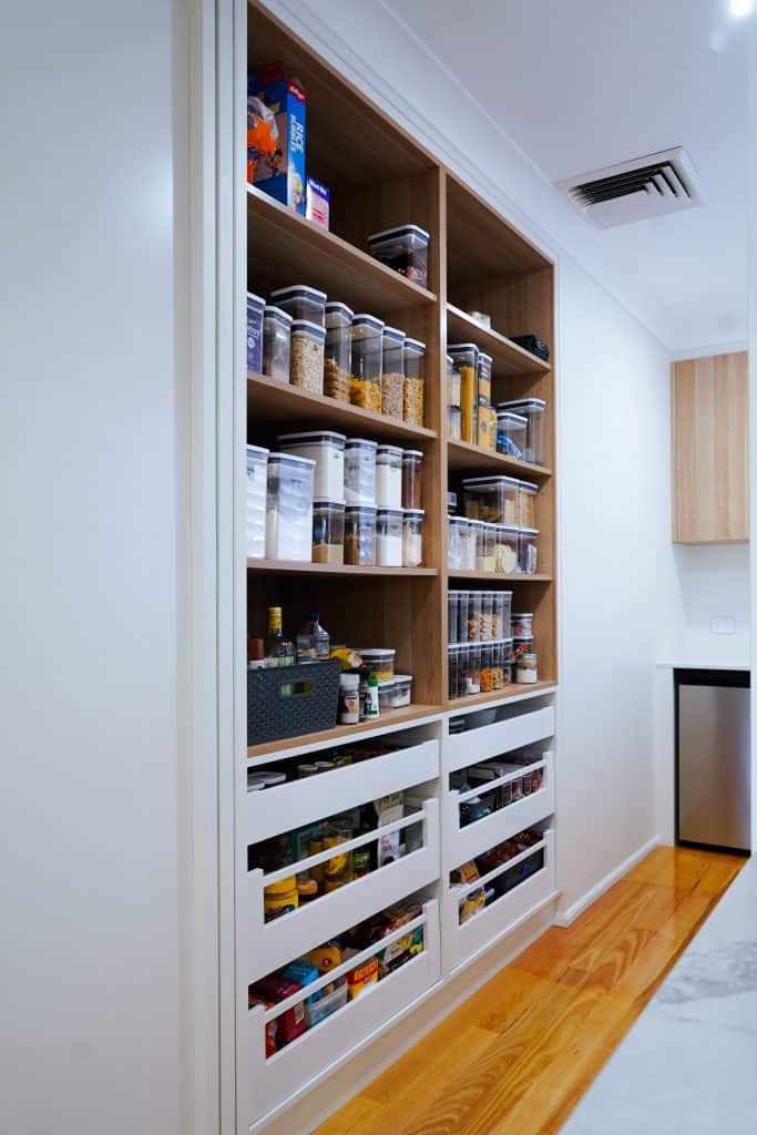 A neatly organised pantry with labelled containers of cereals, pasta, and snacks on wooden shelves, and pull-out drawers filled with various food items in a modern kitchen.