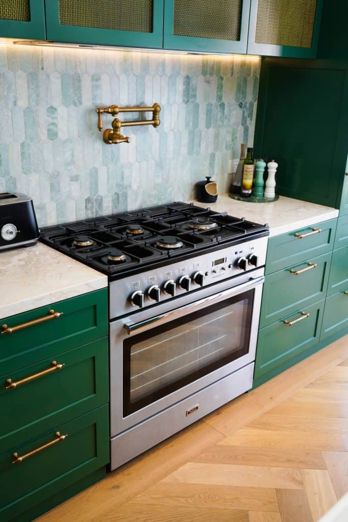 A modern kitchen with green cupboards, gold fittings, a stainless steel gas cooker, marble worktop, and a wall-mounted brass pot filler above a patterned tile splashback. Cooking essentials are neatly arranged nearby.