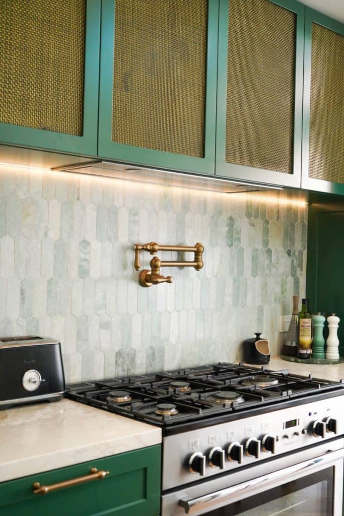 A modern kitchen features green cupboards with brass mesh panels, a marble worktop, a stainless steel gas cooker, a brass pot filler, a tiled splashback, and small appliances and condiments on the counter.