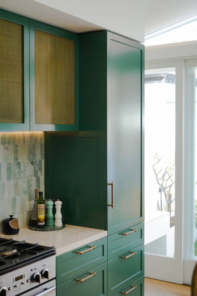 A modern kitchen featuring dark green cupboards with brass handles, a light marble worktop, a patterned green tiled splashback, and a cooker with olive oil and condiments nearby. Natural light streams in from large windows.