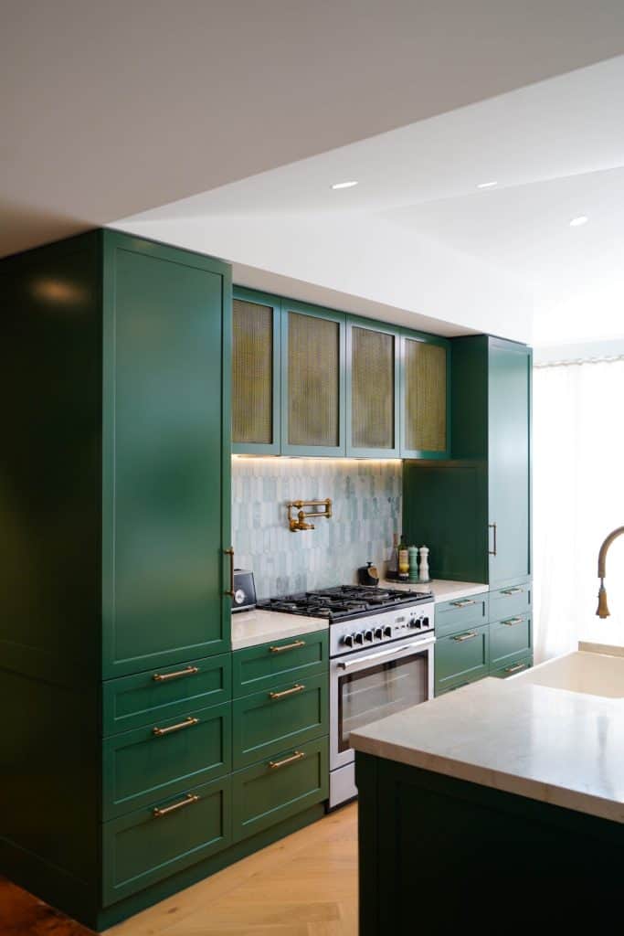 A modern kitchen features dark green cupboards, brass fittings, a stainless steel cooker with a pot filler, and a light tile splashback, all accented by marble worktops and natural light.