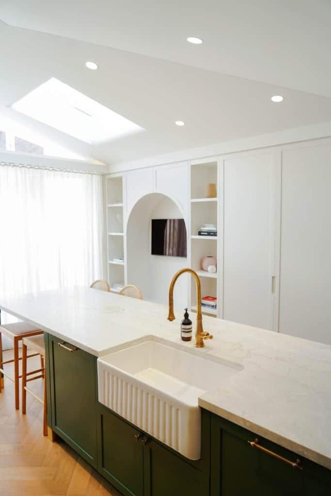 A modern kitchen with a green island, white butler sink, brass tap, marble worktop, and built-in shelves. Natural light streams in from large windows and a skylight above.