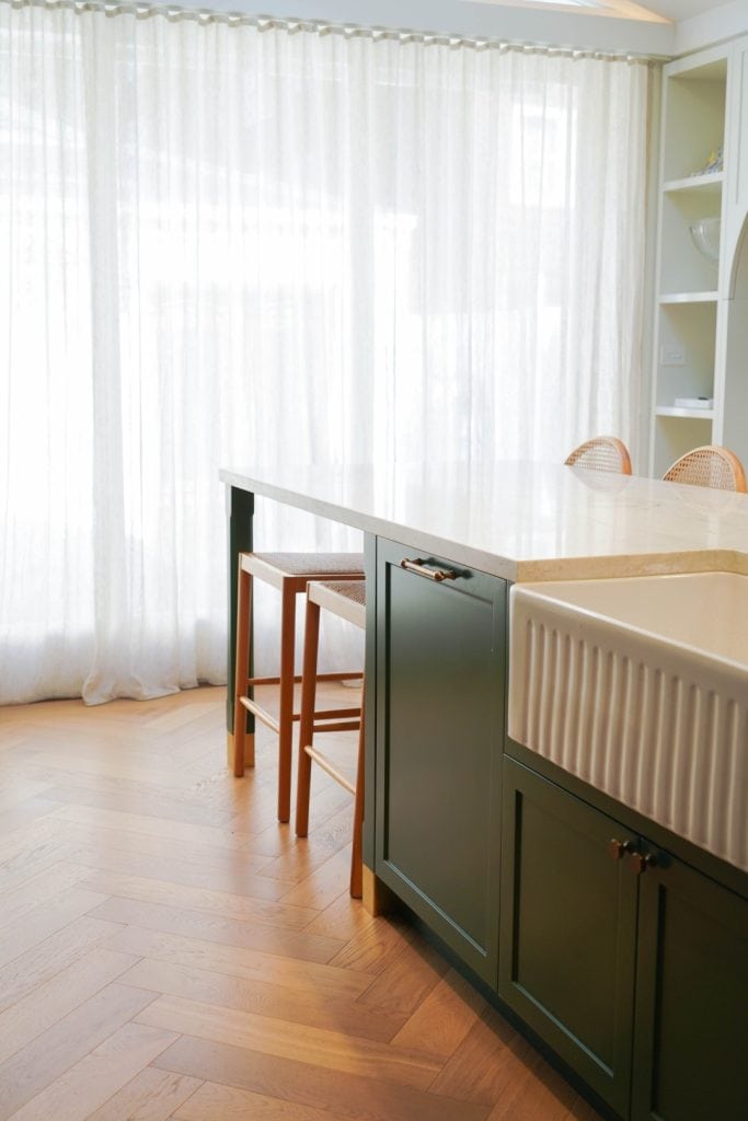 A modern kitchen with a large farmhouse sink, dark green island, white worktop, wooden stools, and herringbone wood floor. Sheer white curtains cover windows in the background, creating a bright, airy feel.