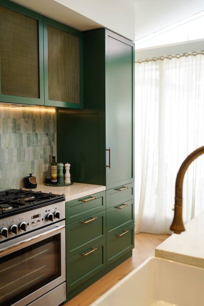 A modern kitchen with dark green cupboards, brass handles, a stainless steel gas cooker, green geometric tile splashback, and sheer white curtains letting in natural light.