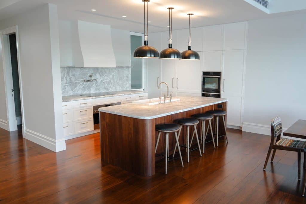 Modern kitchen with a marble island, wooden floors, four stools, three black pendant lights, white cabinetry, and built-in oven. The splashback and worktop are grey marble. Dining table partially visible on the right.