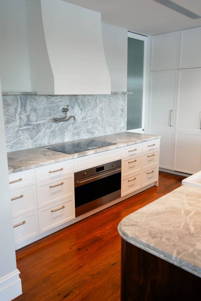 Modern kitchen with white cupboards, marble worktops and splashback, a built-in oven, a hob, and a wall-mounted pot filler tap. The floor is wood, and the overall design is sleek and bright.
