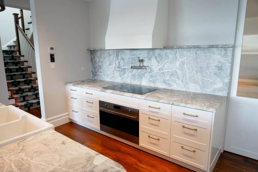 Modern kitchen with white cupboards, marble worktops and splashback, built-in oven, electric hob, pot filler tap above the cooker, and wooden floors. Part of a staircase is visible on the left side.
