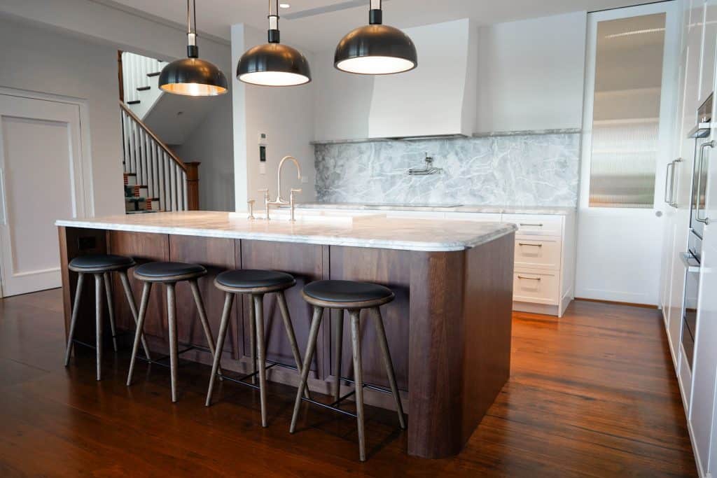 Modern kitchen with a large marble island, four dark wooden stools, pendant lights, white cupboards, stainless steel fittings, and a marble splashback, all on polished wooden floors.