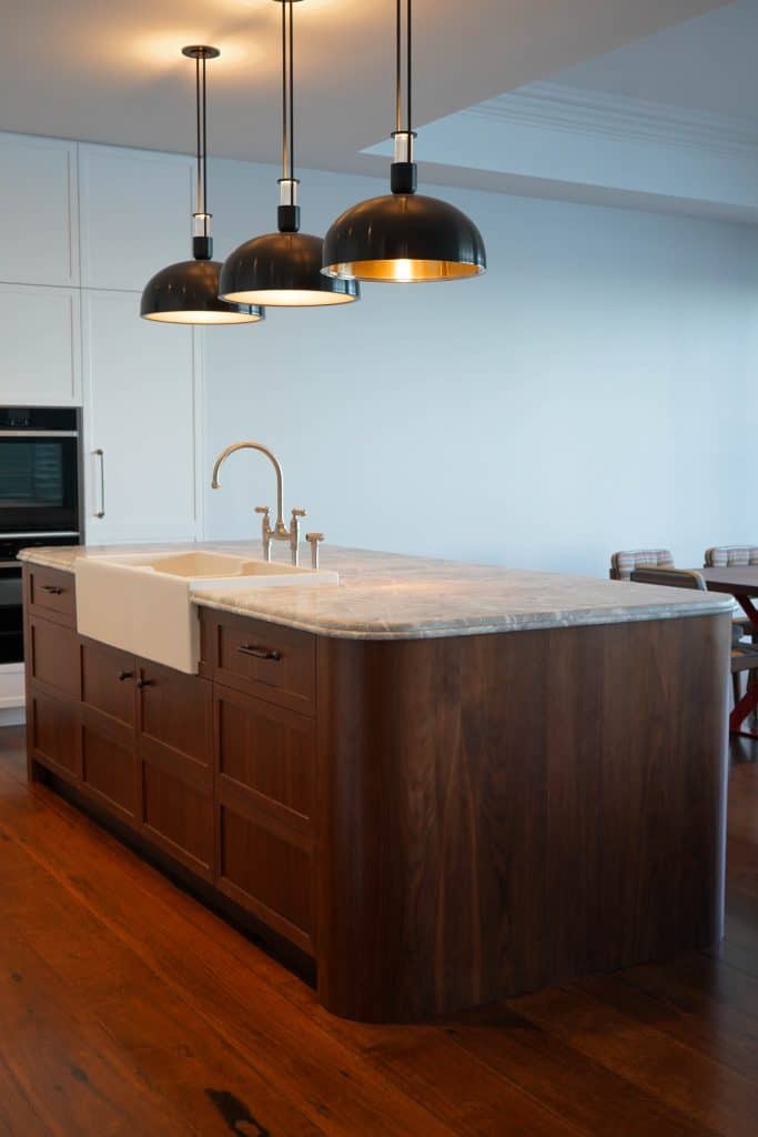 A modern kitchen island with a marble worktop, farmhouse sink, and wooden cupboards, illuminated by three black pendant lights, sits on a hardwood floor in a bright, minimalist space.