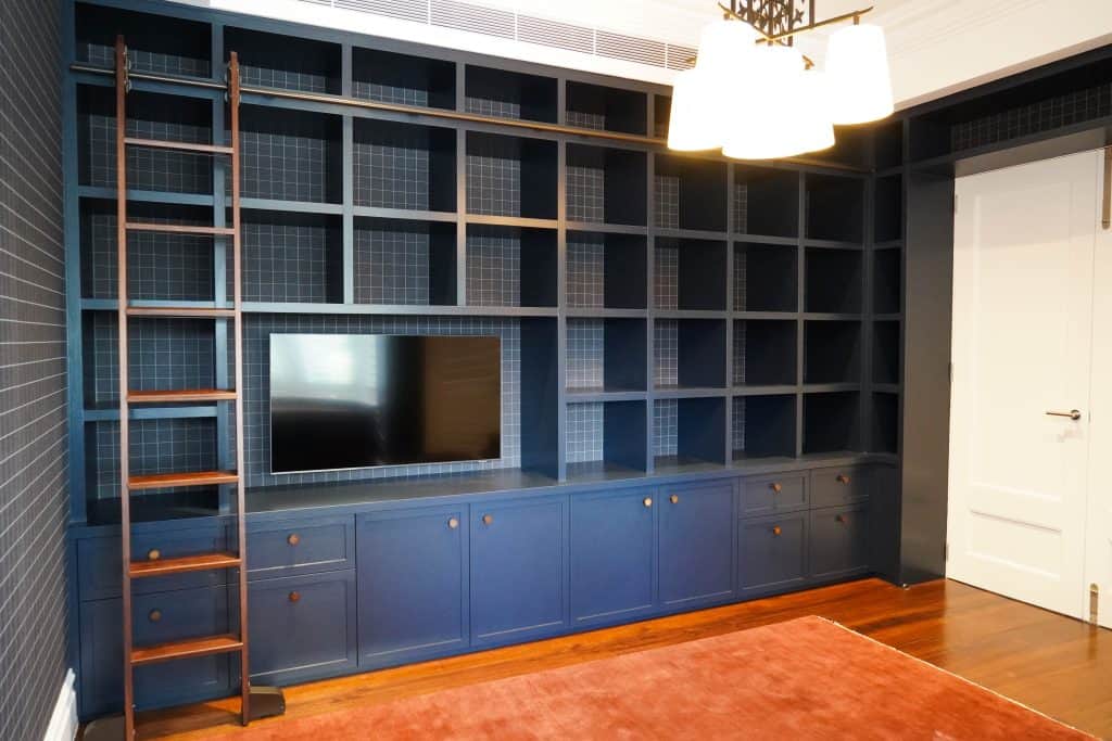 A built-in floor-to-ceiling blue bookcase with empty shelves, cupboards below, a mounted TV, and a wooden ladder on the left. A white door and a reddish-brown rug are also visible in the room.