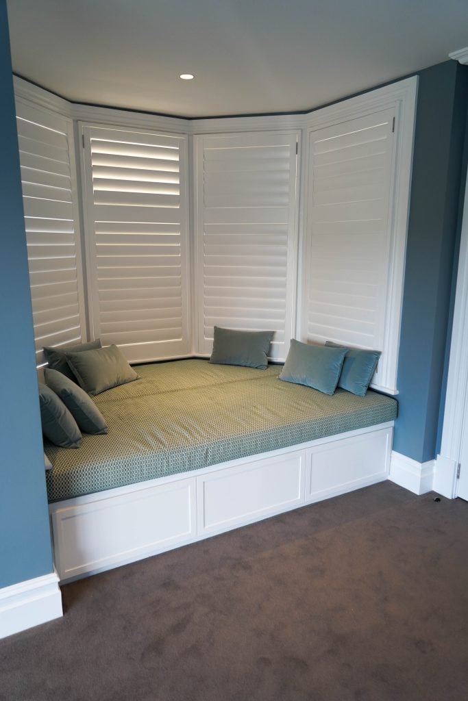 A cosy built-in window seat bed with green cushions and pillows is set in a corner with white shutters and blue walls, above a carpeted floor.