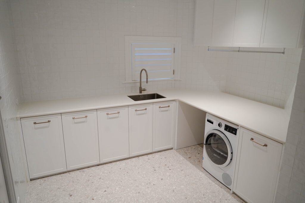 A modern, white utility room with a worktop, sink, cupboards, and a front-loading washing machine under the counter. The room has white tiled walls, terrazzo flooring, and a small window above the sink.