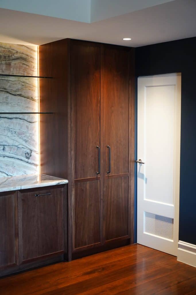 A modern kitchen corner featuring dark wood cupboards, a white door with a vent, marble splashback with subtle lighting, and wooden flooring.